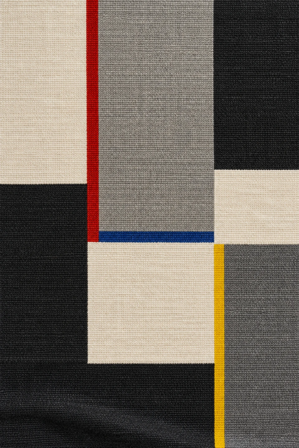 Geometrisches Textil in Creme, Grau und Schwarz mit schmalen roten, blauen und gelben Streifen und einer leichten Falte am unteren Rand.