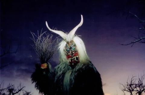 Eine maskierte Figur mit hellem Haar, zwei langen Hörnern und gezeigten Zähnen hält ein Reisigbündel vor dunklem Himmel.