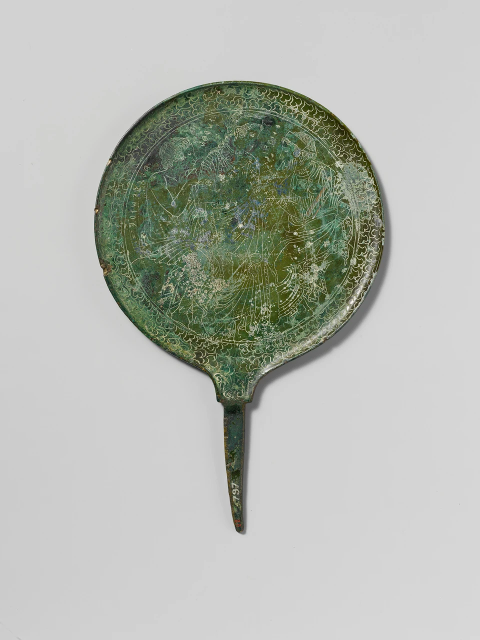 Runder Bronzespiegel mit langem Griff, drei eingeritzten Figuren und ornamentiertem Rand vor grauem Hintergrund; grüne Patina und helle Oberflächenverluste.