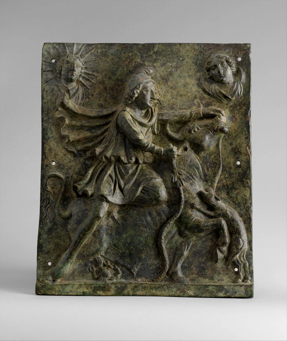 Bronzeplatte mit Mithras über einem Stier, einem Hund rechts unten sowie einer Strahlenbüste links und einer Mondsichelbüste rechts oben. Rand und Befestigungsöffnungen sichtbar.