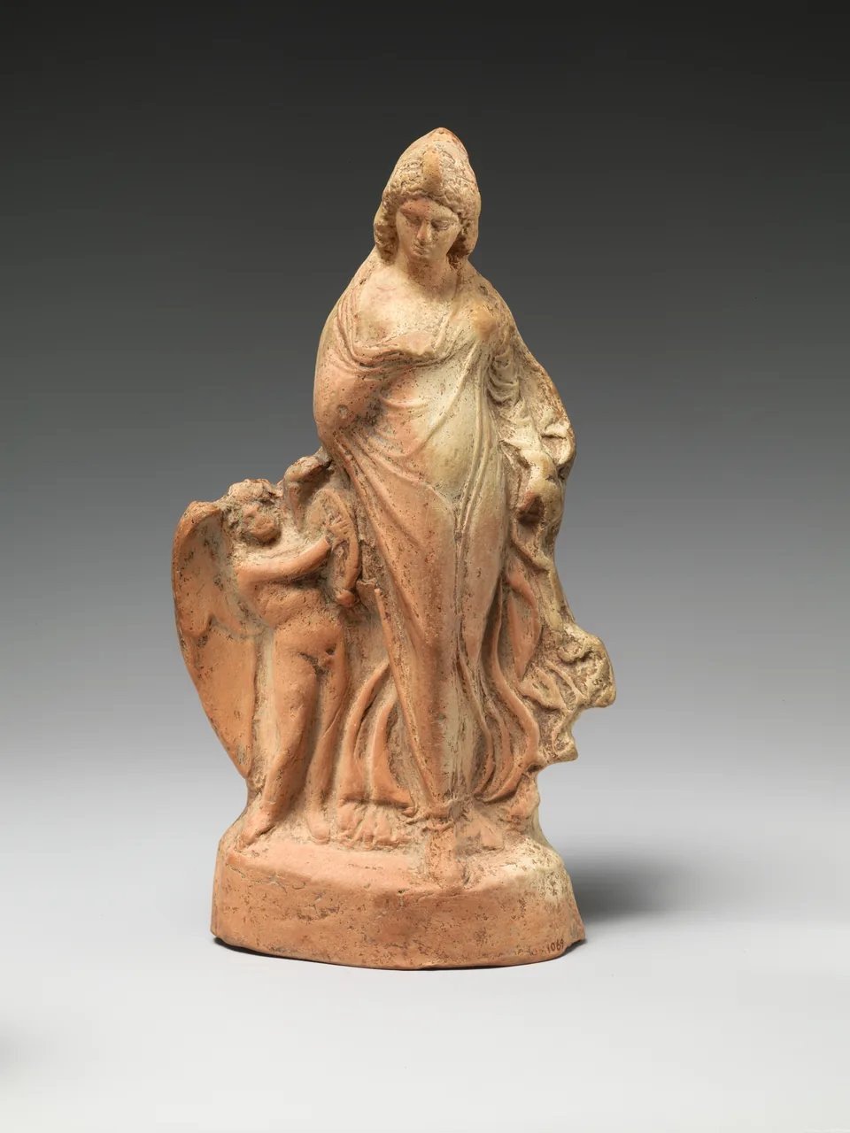 Terrakottafigur einer tanzenden Aphrodite mit kleinem Eros vor hellem Studiohintergrund.