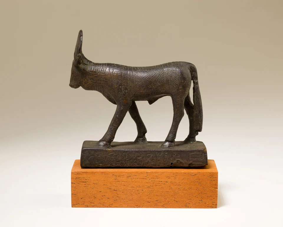 Kleine dunkle Bronzefigur eines schreitenden Stiers auf beschrifteter Basis, aus linker Seitenansicht.