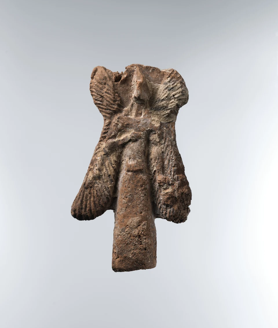 Kleine braune Tonfigur mit vier seitlichen Flügeln, vorspringendem schnabelähnlichem Gesicht und langem Gewand. Ein Arm liegt angewinkelt vor der Brust; seitlich ist ein kleines Gefäß erkennbar.