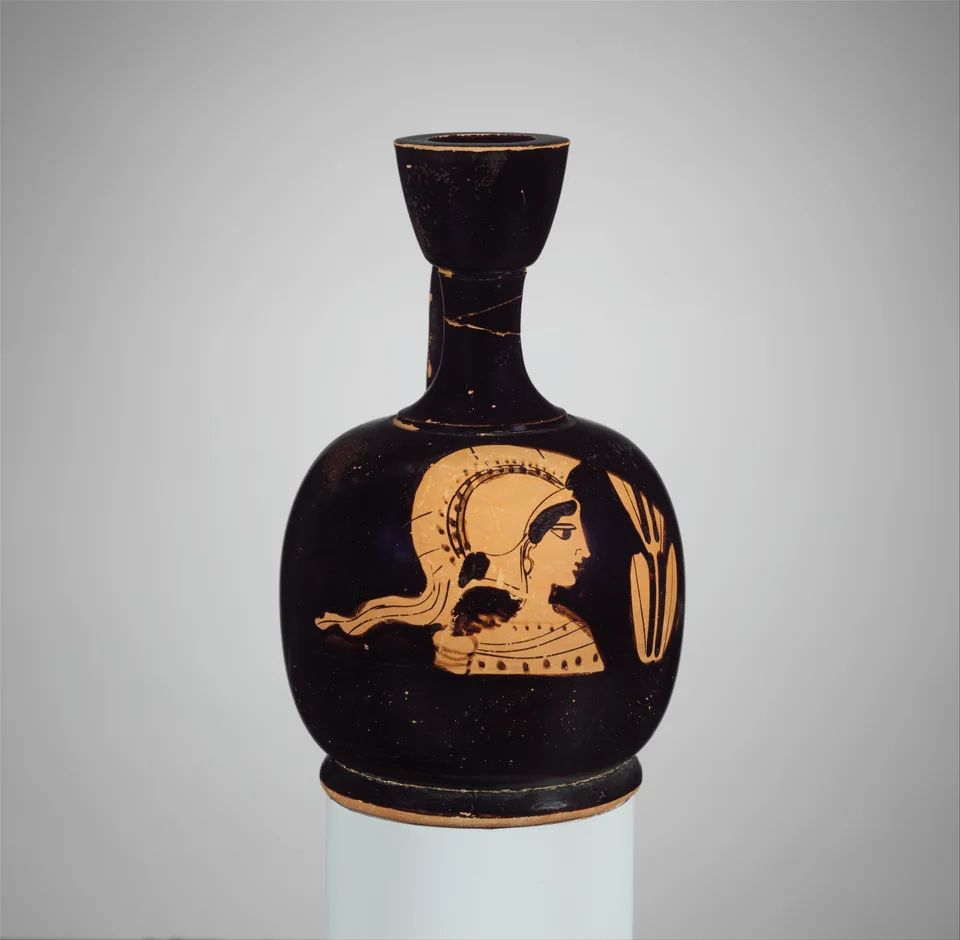 Kleine schwarze attische Ölkännchen mit rotfiguriger Büste der behelmten Athena neben einem Olivenzweig auf hellem Studiohintergrund.