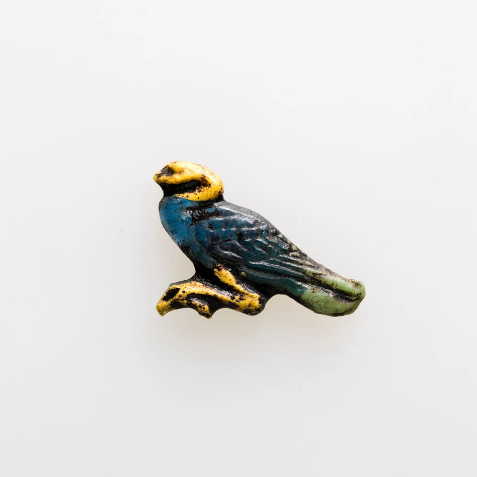 Kleines blau-grünes Glasamulett in Vogelgestalt mit gelben Partien vor weißem Hintergrund.