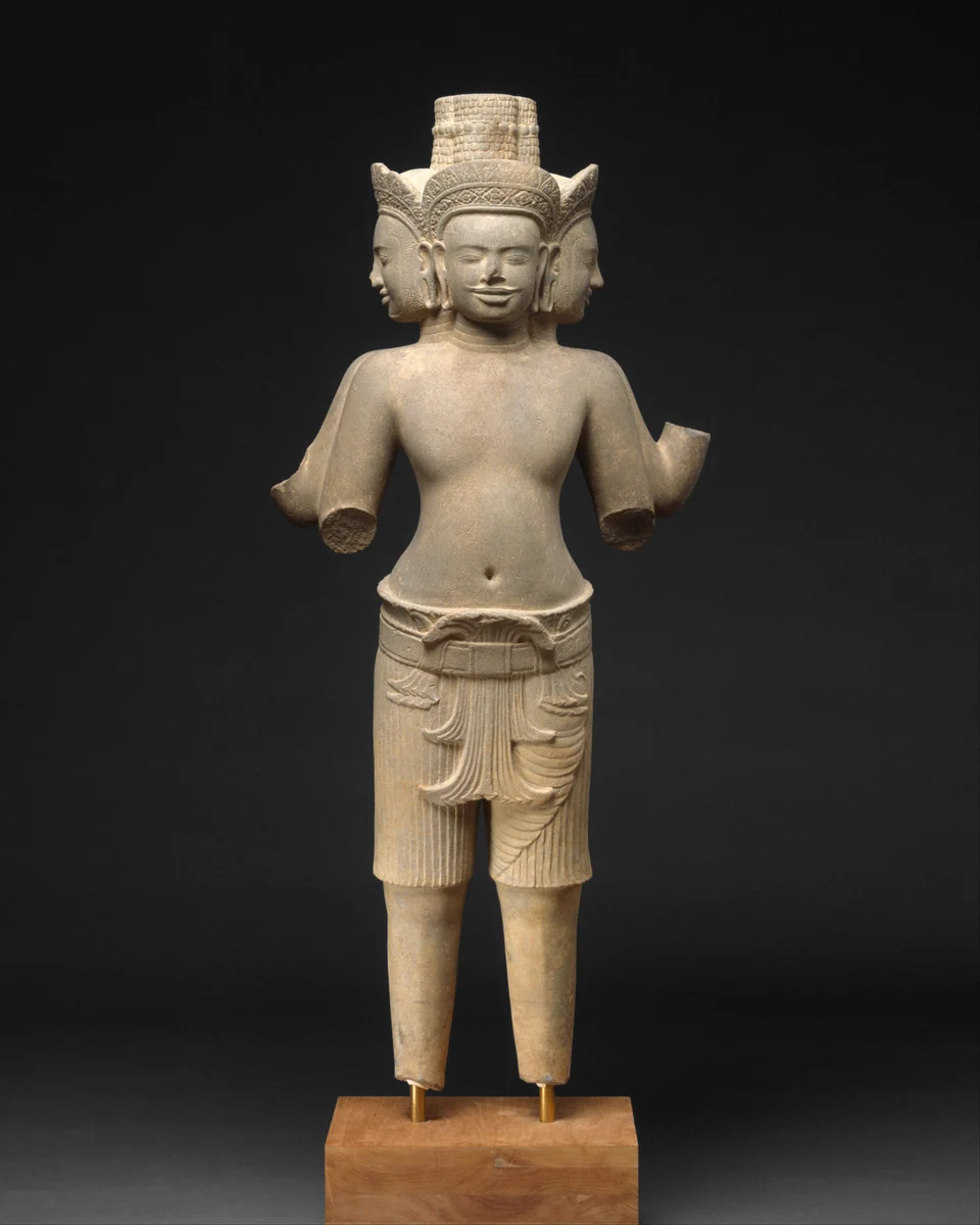 Sandsteinfigur mit einem vorderen und zwei seitlich sichtbaren Gesichtern, hohem Kopfschmuck und abgebrochenen Armen; die Beine stehen auf einer modernen Halterung.