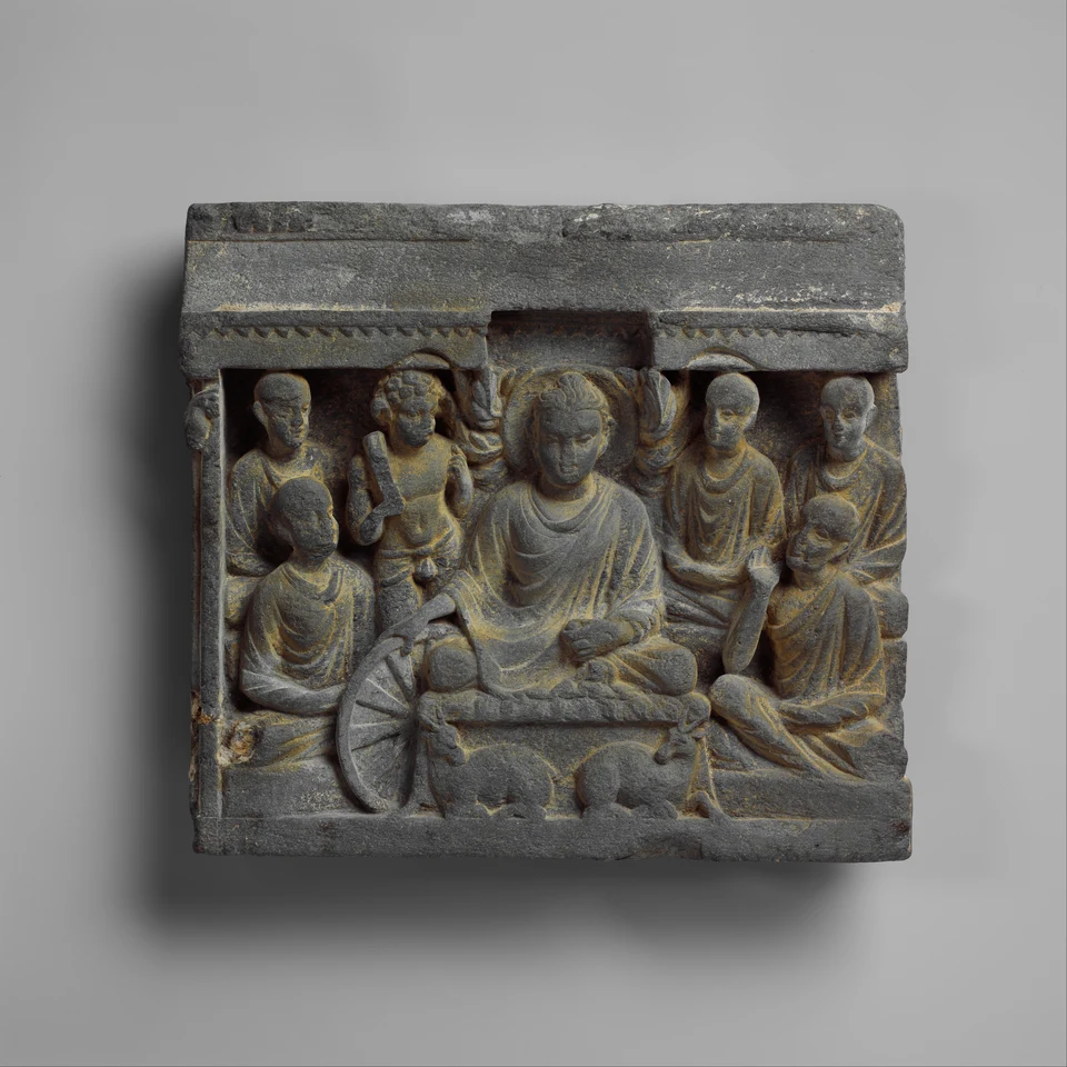 Graues Steinrelief: Ein Buddha mit Nimbus sitzt inmitten mehrerer stehender Figuren; seine Hand reicht zu einem Rad. Eine muskulöse Figur hält einen dicken Stab.