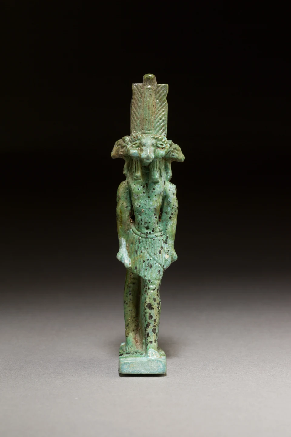 Grünliches Fayenceamulett mit einer menschengestaltigen Figur und mehreren seitlich ausgerichteten Widderköpfen vor dunklem Hintergrund.