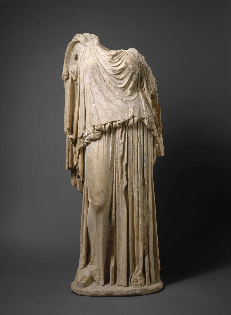 Kopf- und armloser Torso einer römischen Marmorstatue der Eirene vor dunklem Hintergrund; das helle Gewand fällt in tiefen Falten bis zum Sockel.