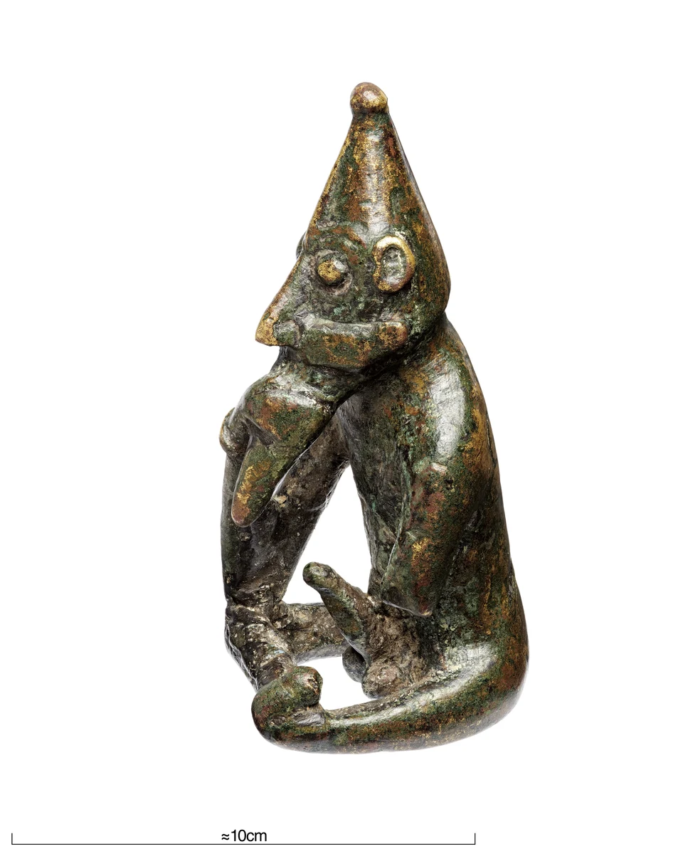 Sitzende männliche Metallfigur mit spitzer Kopfbedeckung, großen Augen und rechter Hand am Bart. Der Körper ist nackt, der Phallus aufgerichtet und der linke Arm abgebrochen. Unten ein Maßstabsbalken.
