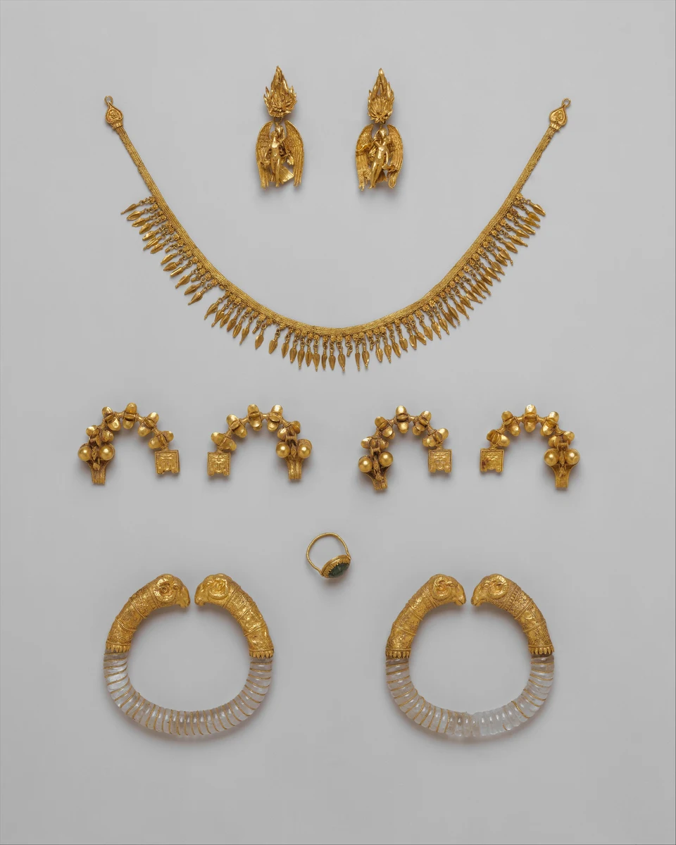 Schmuckensemble auf grauem Grund: oben zwei goldene Anhänger mit Knaben zwischen Adlerflügeln, darunter Halskette, vier Fibeln, Ring und zwei Armreifen mit goldenen Tierköpfen.