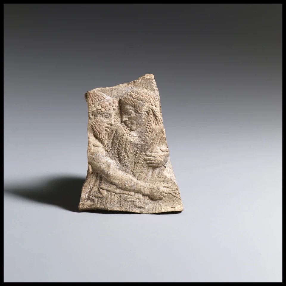 Terrakottafragment mit zwei nahen Oberkörpern. Links eine bärtige Gestalt; ein Arm greift quer vor die andere Figur. Oben und seitlich verlaufen Bruchkanten.