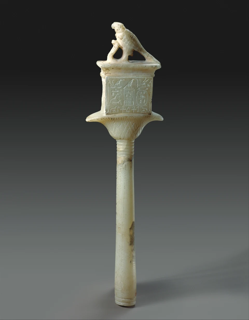 Helles steinernes Sistrum mit schmalem Stiel, Inschriftfeld und kleinem Falken an der Spitze.
