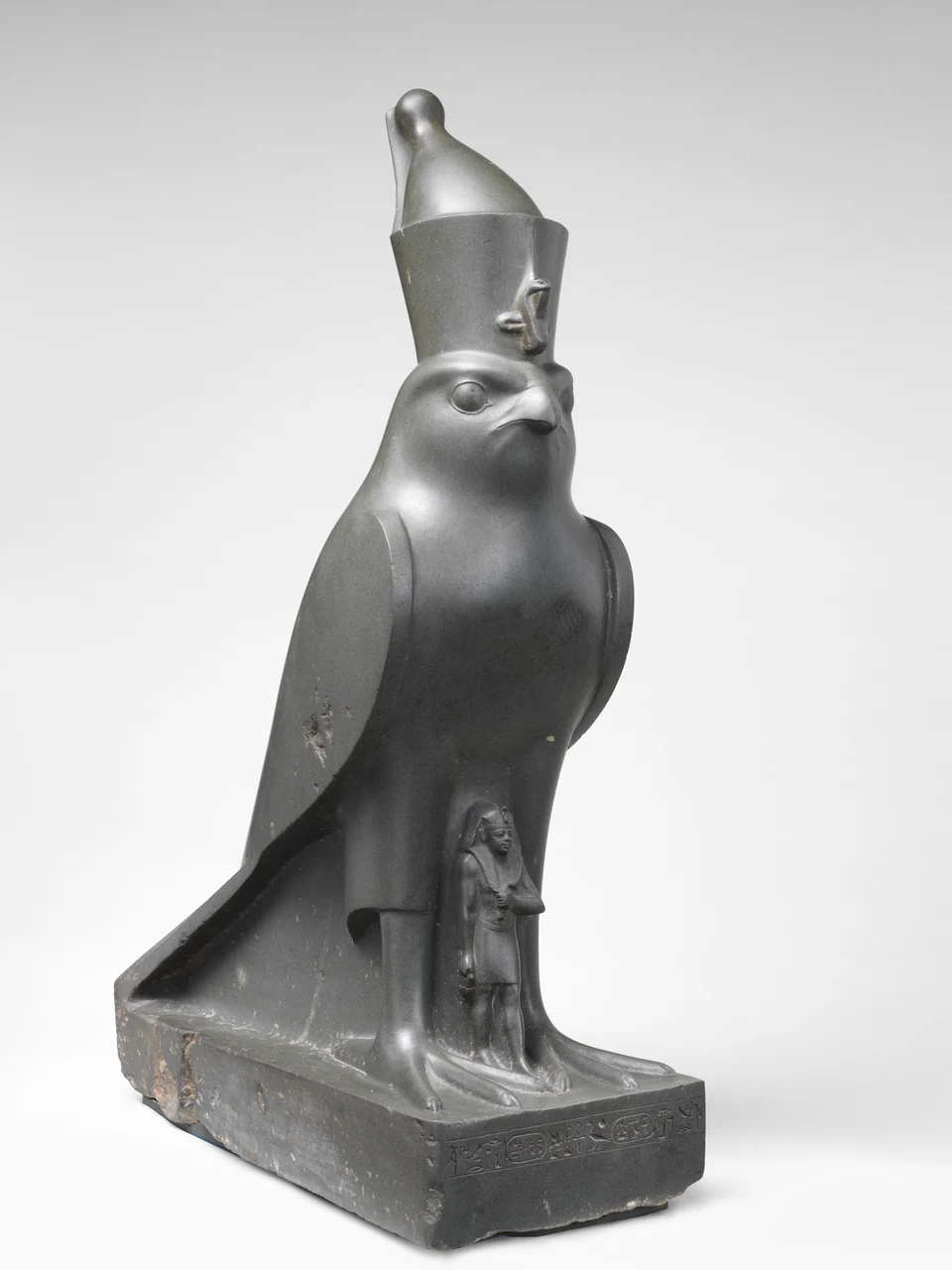 Große dunkle Falkenstatue mit hoher Doppelkrone; zwischen ihren Beinen steht eine sehr kleine Königsfigur.