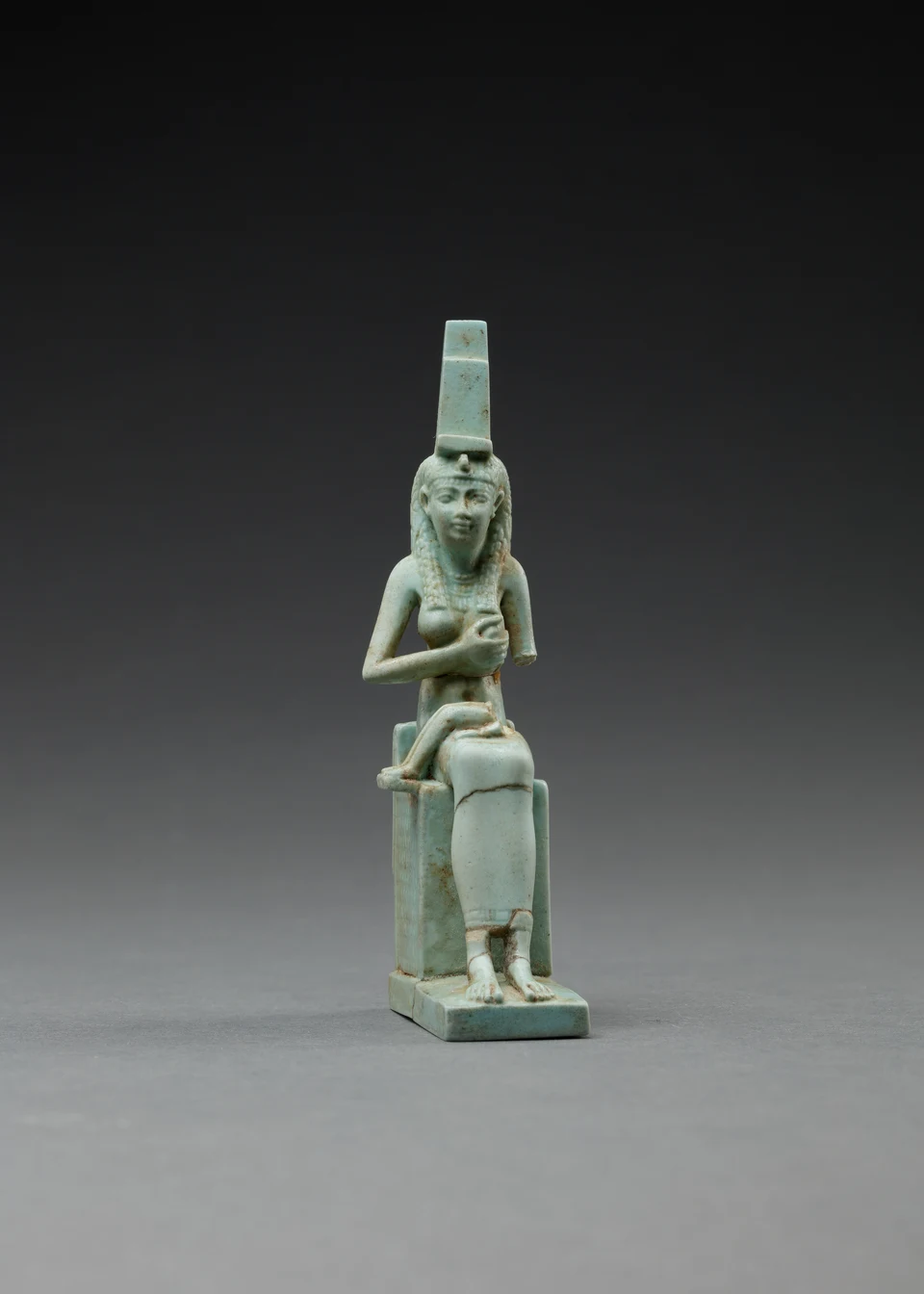 Türkisfarbene, beschädigte Figur einer sitzenden Frau mit hohem Thronzeichen auf dem Kopf. Auf ihrem Schoß liegen erhaltene Beine einer kleineren Figur; deren Oberkörper fehlt.