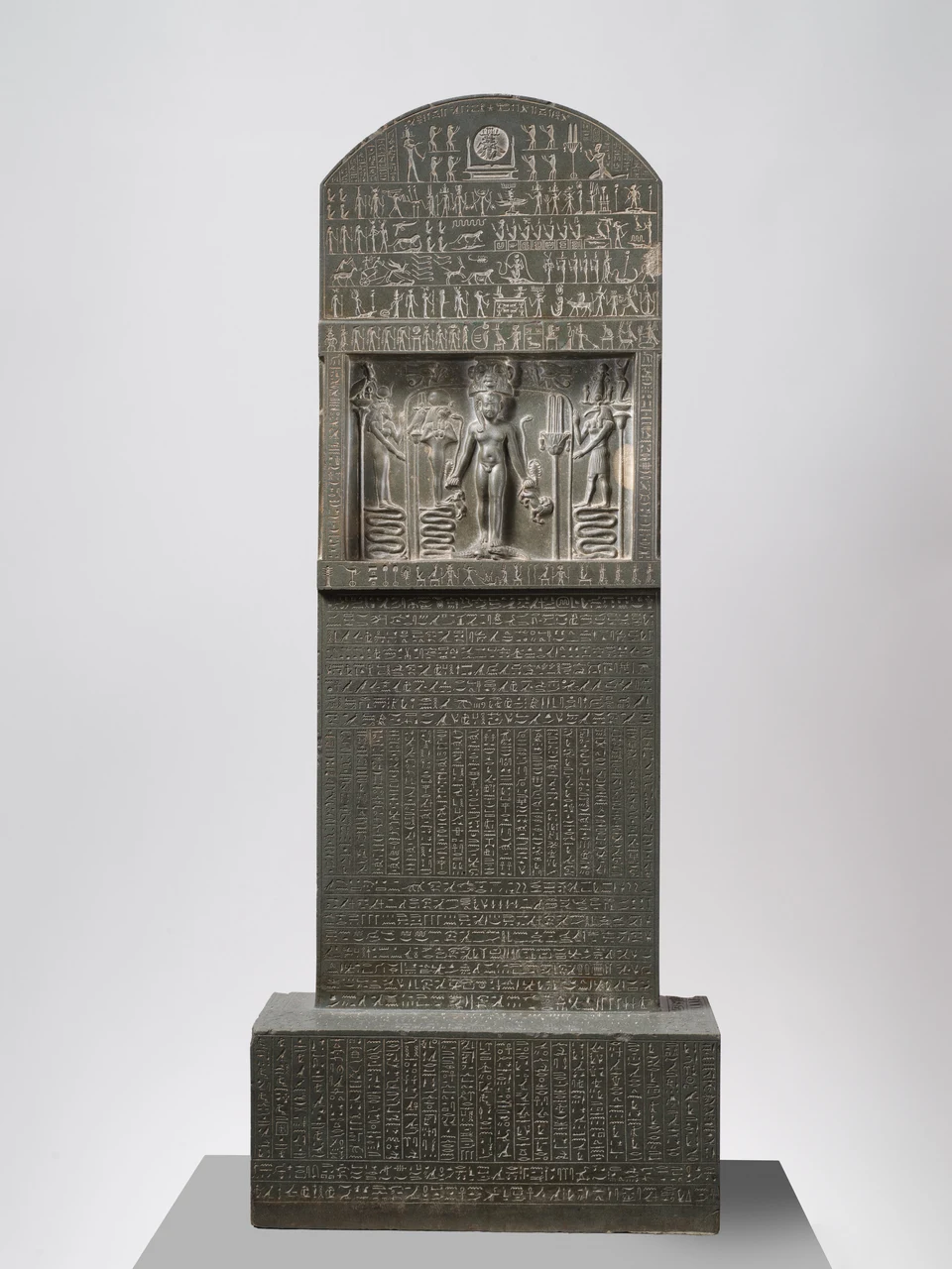 Hohe dunkle Stele mit gerundetem oberen Abschluss, dicht angeordneten Figuren und Schriftfeldern. In der Mitte steht eine kleine frontale Gestalt über zwei Krokodilen.