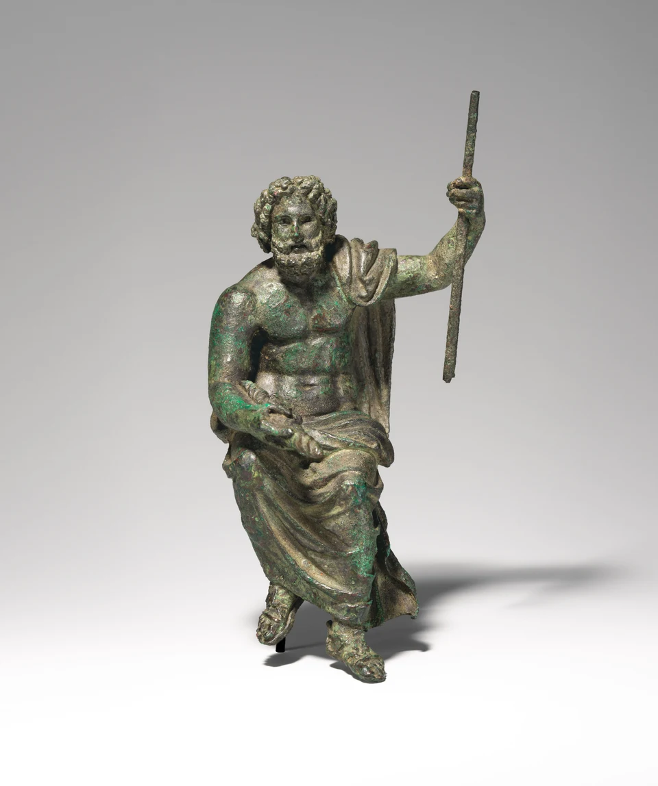 Kleine grünbraun patinierte Bronzefigur eines bärtigen sitzenden Mannes, mit Gewand über Schulter und Beinen, langem Stab in der erhobenen linken Hand und Blitzbündel in der rechten.