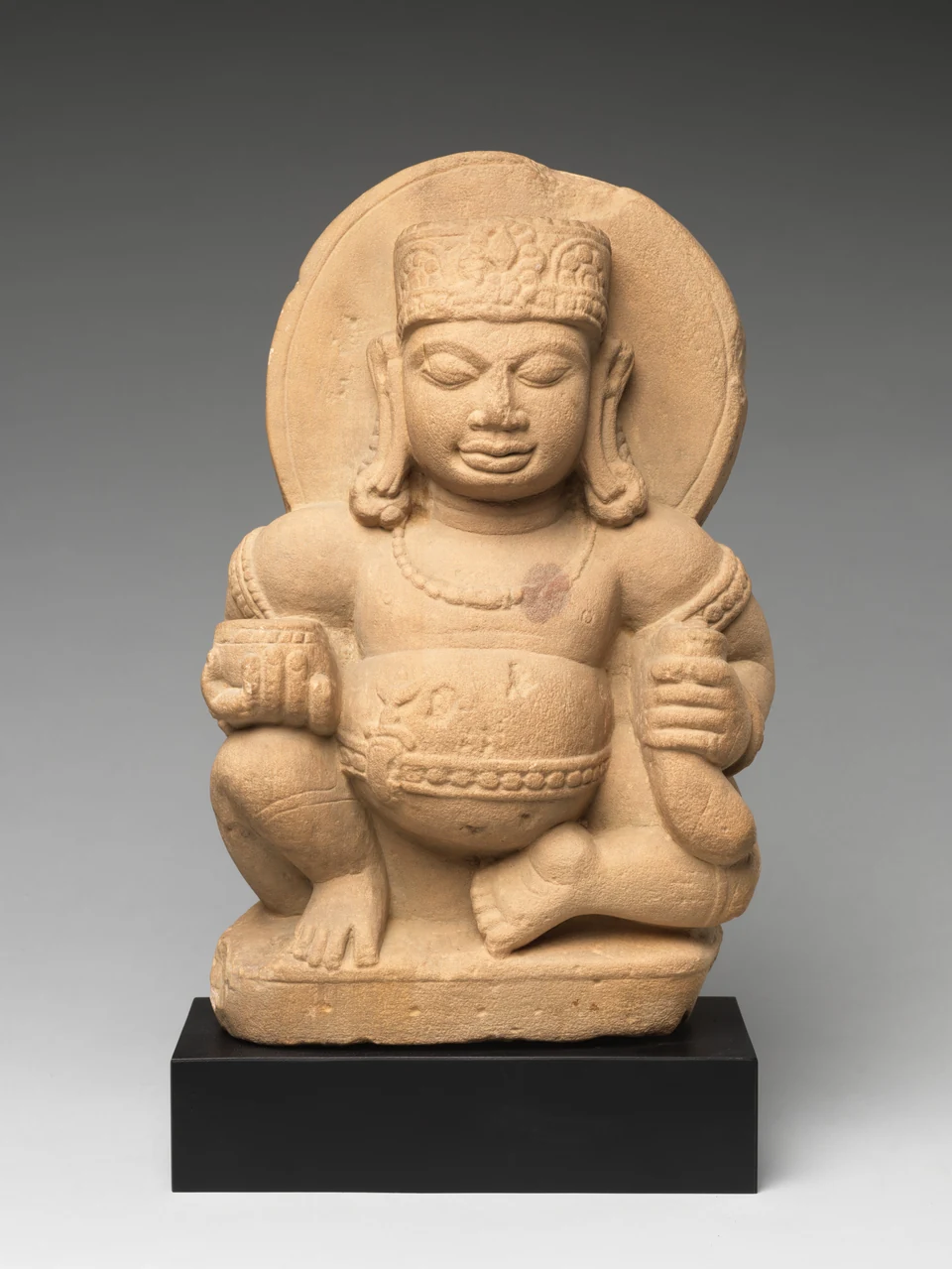 Sitzende rundbäuchige Steinfigur mit Kopfschmuck, rundem Hintergrund, Becher und länglichem Gegenstand; schwarzer Museumssockel bleibt sichtbar.