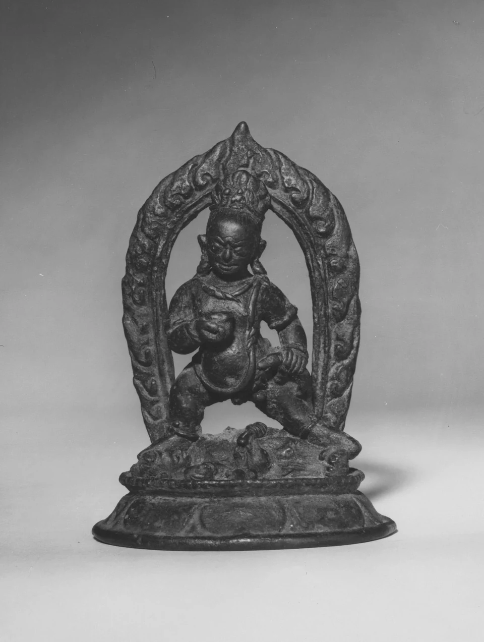 Schwarzweiße Museumsaufnahme einer kleinen Bronzefigur unter einem ornamentierten Bogen auf einem mehrstufigen Sockel.