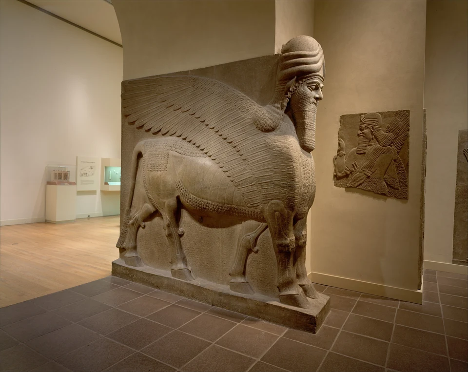 Ein steinerner geflügelter Stier mit bärtigem Menschenkopf und Hörnerkappe steht im Museum. Die schräge Ansicht zeigt mehrere Hufbeine, einen großen gefiederten Flügel und Schriftflächen am Körper.