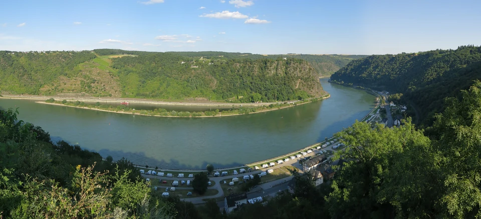 Blick über eine Rheinschleife zum bewaldeten Loreley-Felsen; am Ufer Straßen, Bahn und ein Campingplatz.