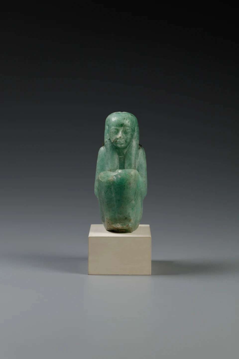 Kleine grünliche Feldspatfigur der Maat auf einem Sockel vor dunklem Hintergrund; eine Feder ist im Frontalfoto nicht sicher erkennbar.