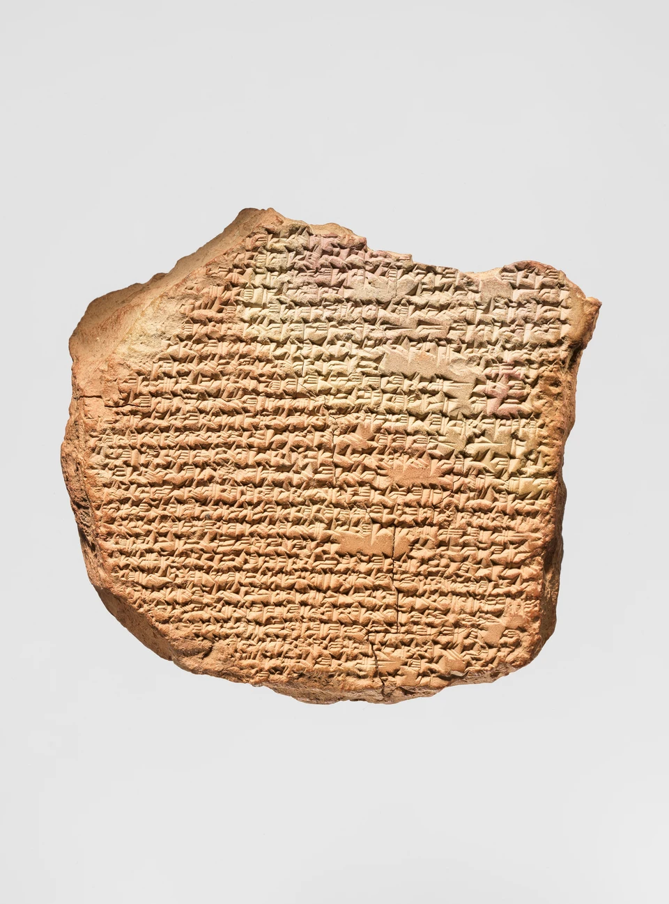 Braunes Tonfragment auf hellgrauem Grund mit dicht gesetzten Keilzeichen, unregelmäßig gebrochener Oberkante und hellen beschädigten Stellen zwischen den Zeilen.