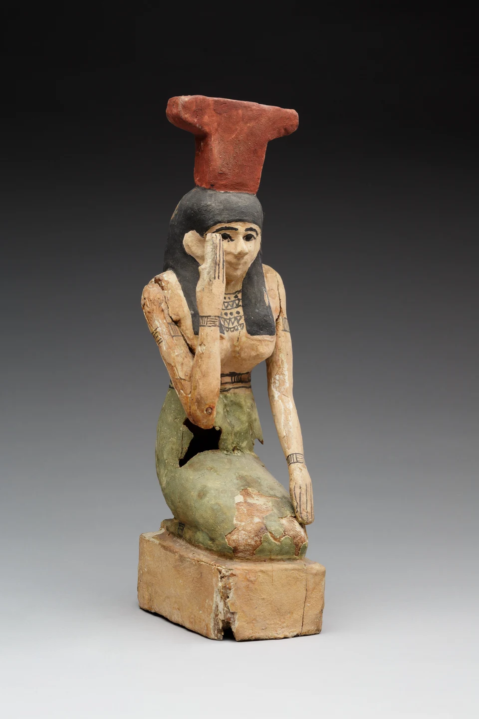 Bemalte kniende Holzfigur der Nephthys mit rotem Namenszeichen auf dem Kopf und einer Hand am Gesicht.
