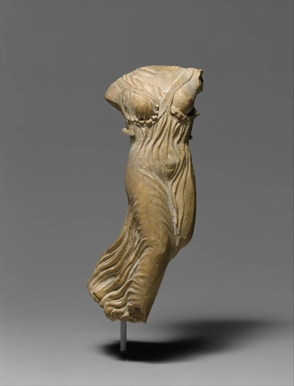 Fragment einer hellen griechischen Terrakottafigur vor grauem Hintergrund: der Kopf und die Arme fehlen, das Gewand ist in bewegten Falten modelliert; rückwärtige Flügel sind nicht erhalten.