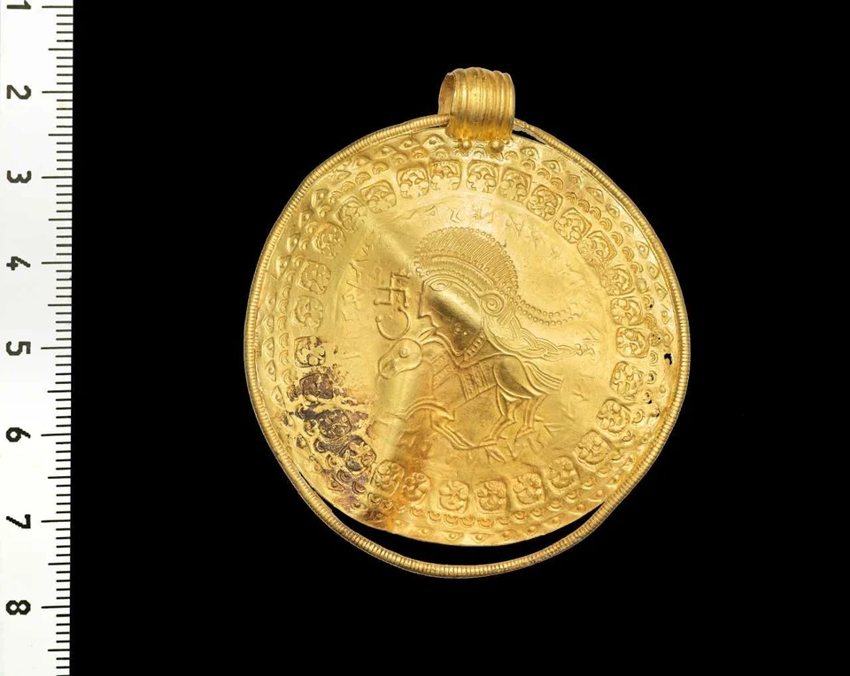 Goldener Anhänger auf schwarzem Grund mit Maßstab links: ein großer Profilkopf über einem vierbeinigen Tier, umlaufende Zeichen und Ornamente, Öse oben und dunkle Beläge links unten. Vor dem Gesicht steht ein antikes Hakenkreuz.
