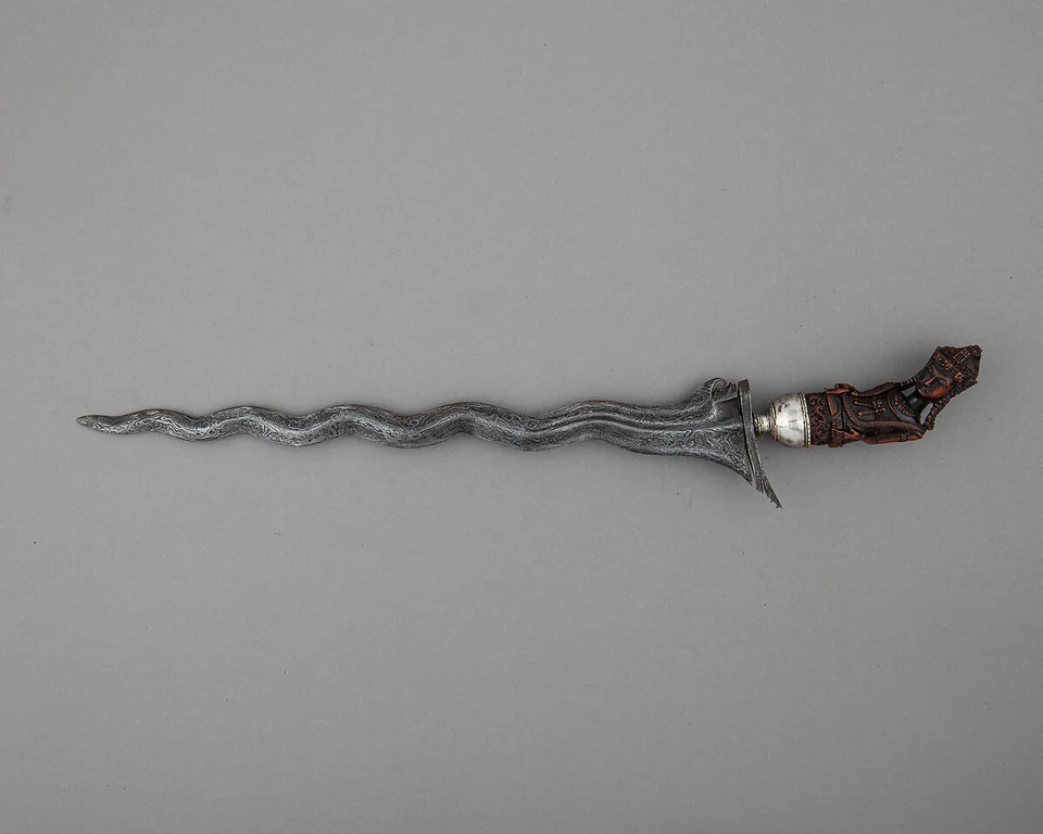Ein vollständiger Kris mit gewellter Metallklinge und geschnitztem Figurengriff liegt waagerecht auf grauem Grund.
