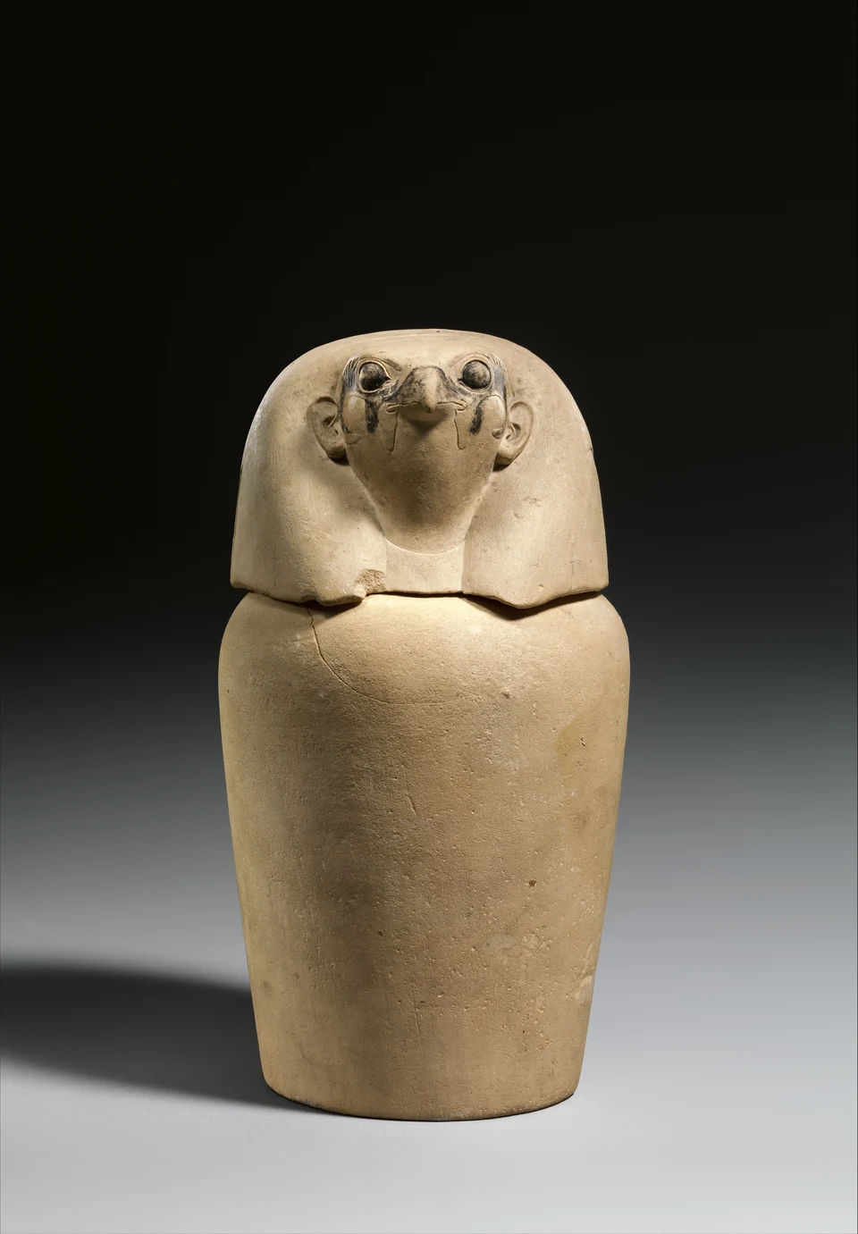 Heller Kanopenkrug mit falkenköpfigem Deckel vor dunklem Hintergrund; auf dem Kopf sind Augen und Schnabel erkennbar.