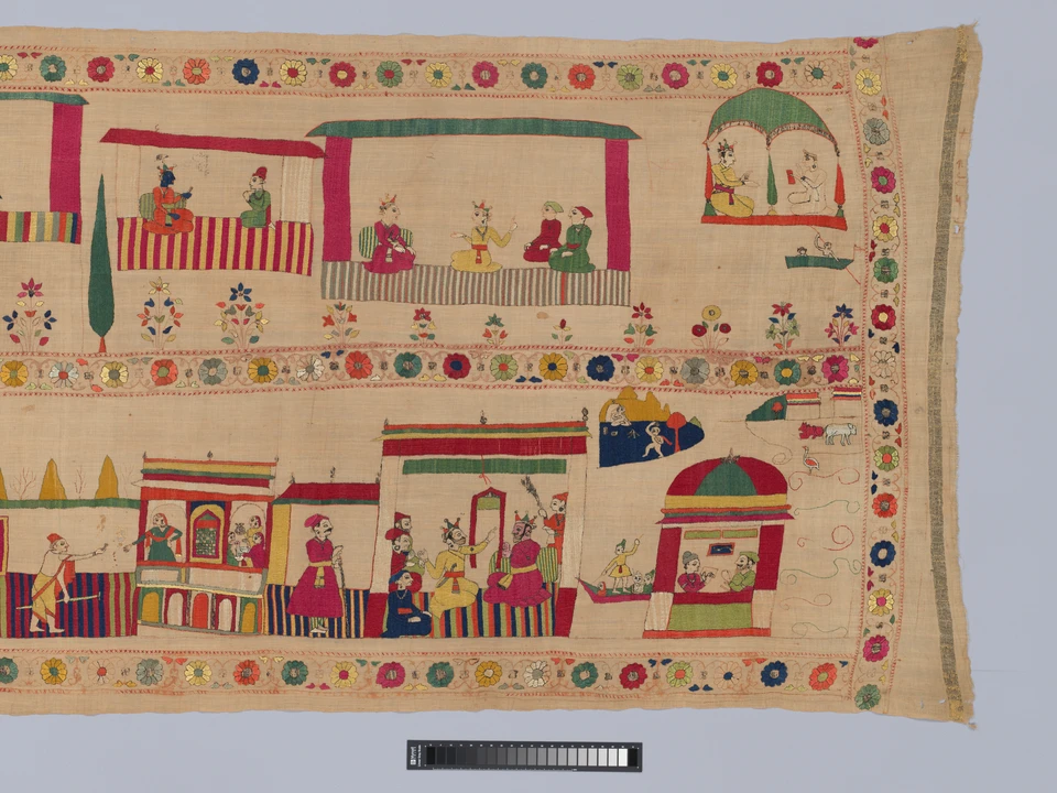 Teilansicht einer langen Stickerei: zwei Reihen mit kleinen Figuren und Gebäuden werden von bunten Blumenbändern eingefasst; am rechten Rand endet das Textil.