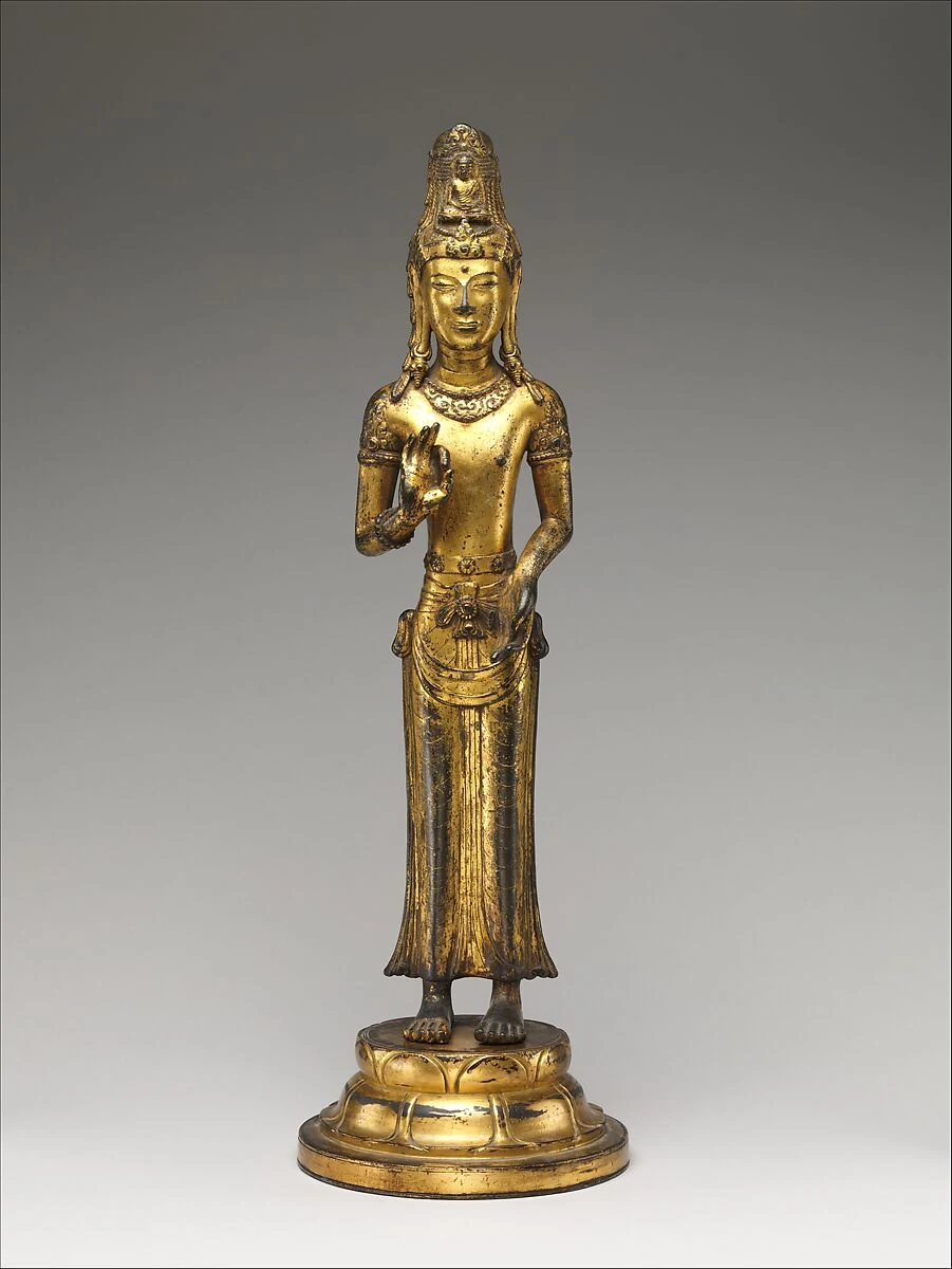 Schlanke, goldfarbene stehende Figur mit hohem Kopfschmuck, einer vor der Brust erhobenen Hand und langem Gewand auf einem Lotusfuß.