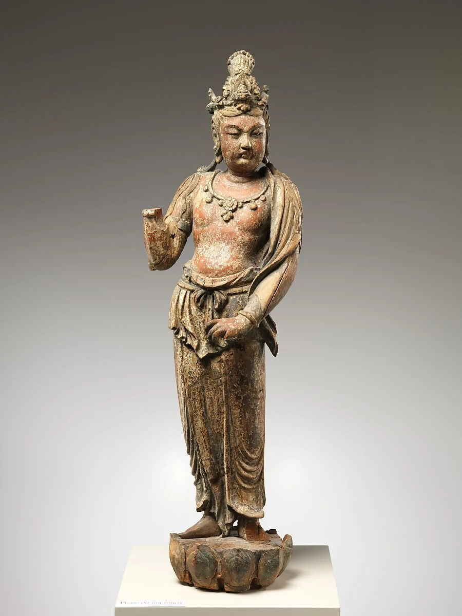 Stehende Holzfigur mit verblassten Farbspuren, hohem Kopfschmuck und drapiertem Gewand; an einem erhobenen Arm fehlt die Hand.