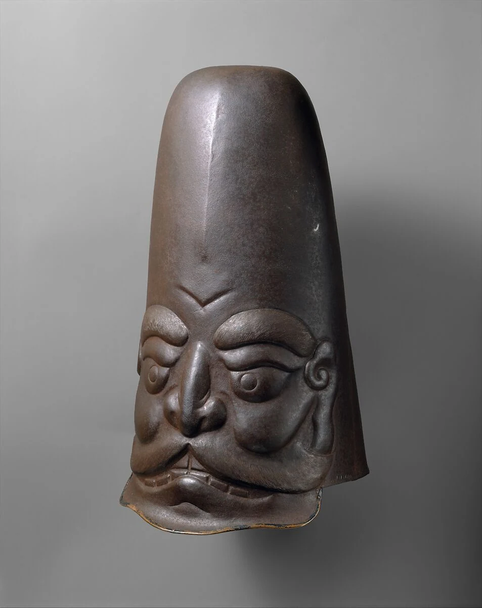 Ein dunkler hoher Helm ist als Gesicht mit großen Augen, gebogenen Brauen und hohem Schädel geformt.