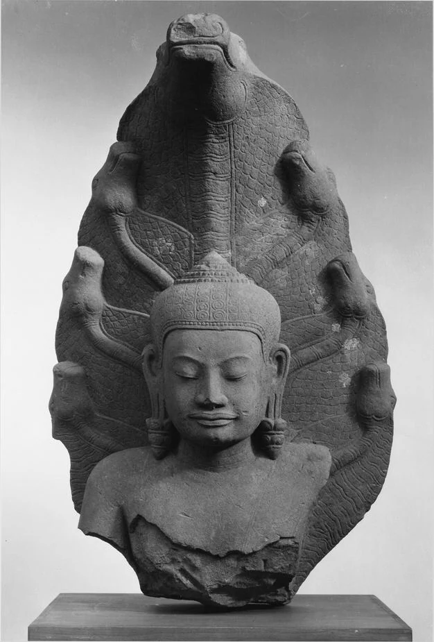 Frontalansicht eines gebrochenen Buddha-Oberkörpers und einer Haube mit mehreren Schlangenköpfen vor hellem Hintergrund.