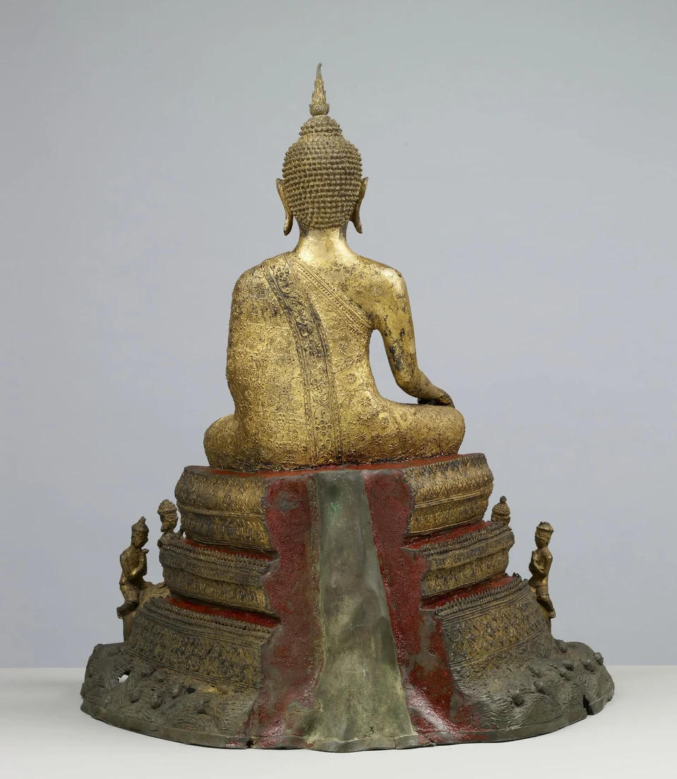 Rückansicht einer vergoldeten sitzenden Buddhafigur auf einem gestuften Sockel mit kleineren Figuren im unteren Bereich.