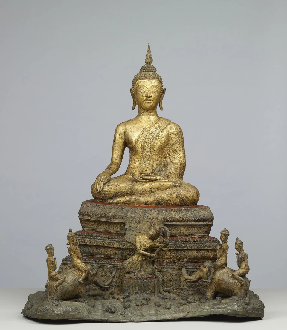 Frontansicht einer vergoldeten sitzenden Buddhafigur auf einem gestuften Sockel mit kleineren Figuren im unteren Bereich.