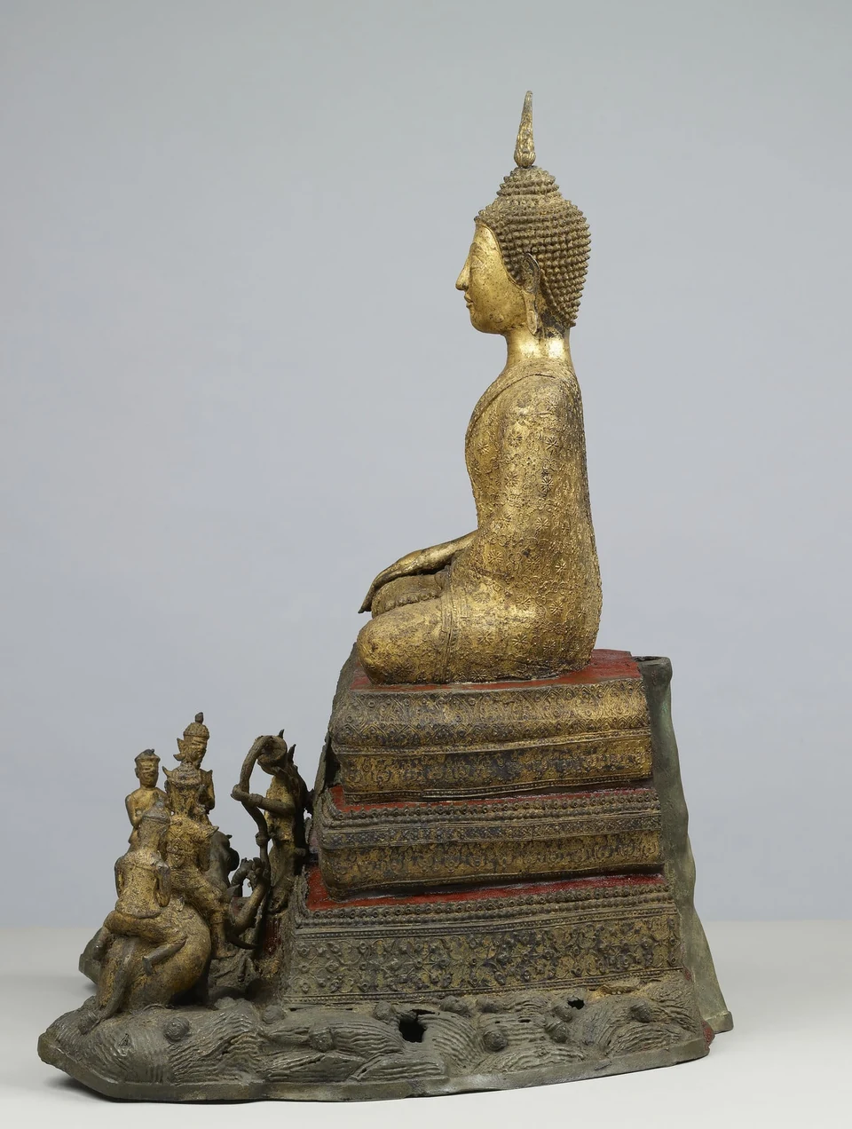 Linke Seitenansicht einer vergoldeten sitzenden Buddhafigur auf einem gestuften Sockel mit kleineren Figuren im unteren Bereich.