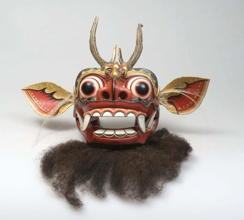 Rote Maske mit runden Augen, breiten Ohren, hohen Fortsätzen, weißen Zähnen und dunklem Bart vor hellem Hintergrund.