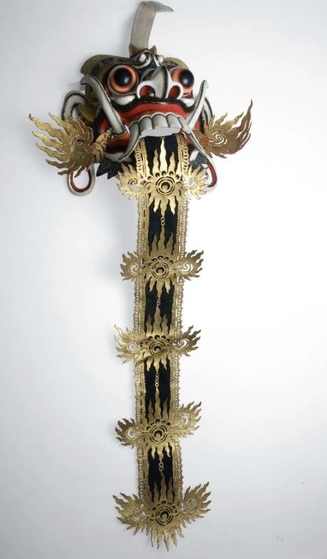 Bemalte Maske mit runden Augen, weißen Zähnen und einer langen schwarz-goldenen Zunge vor hellem Hintergrund.