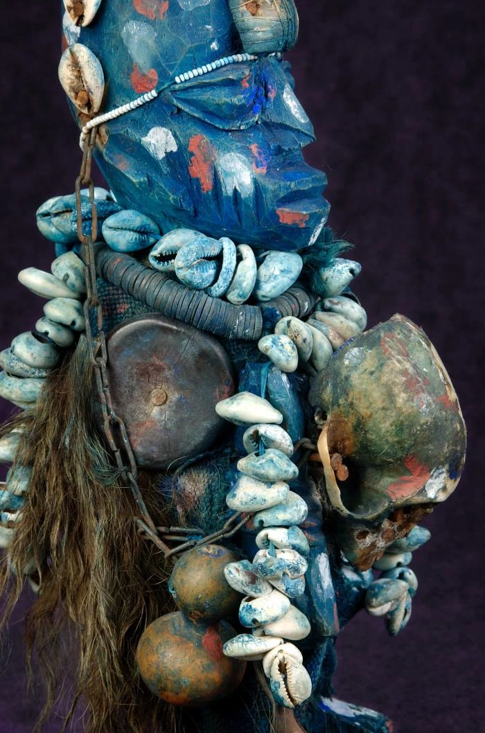 Detailfoto einer blau bemalten geschnitzten Shapona-Figur mit Kaurimuschelketten und angehängten Gegenständen.