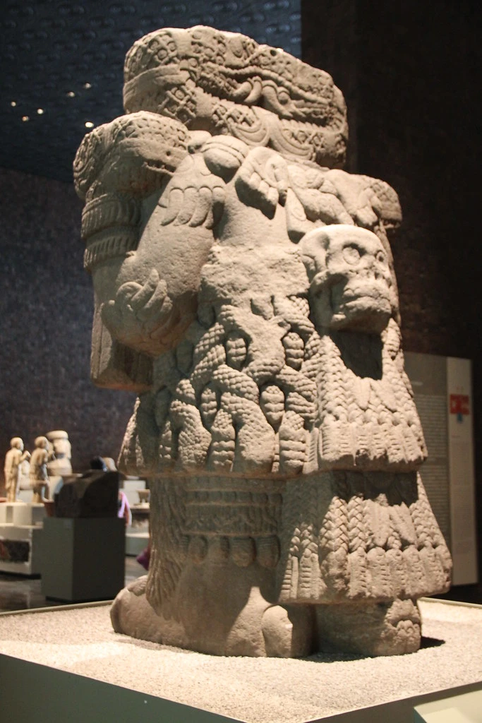 Tatsächliche Steinskulptur schräg von vorn: hohe Gestalt mit Schlangenformen und zentralem vorspringendem Schädel.