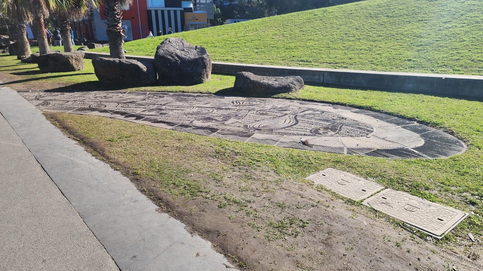 Geschwungene flache Fläche mit dichtem Linienmuster neben dunklen Steinen in einer Grünanlage; ein größerer Stein trägt Gravuren, dahinter liegen ein Rasenhang und ein Gebäude.