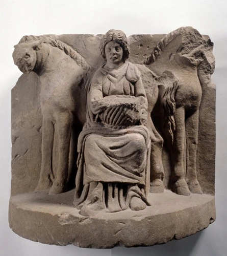 Steinrelief mit sitzender Frau zwischen zwei Pferden und einem Korb vor dem Körper; beschädigter Rand und Steinoberfläche bleiben sichtbar.