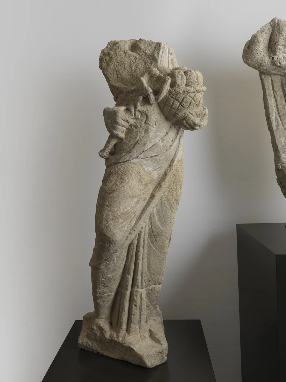 Kopflose Steinfigur Rosmertas aus Paris mit Fruchtkorb, Gewandfalten und fragmentiertem Attribut; moderne Museumsaufstellung vor heller Wand.