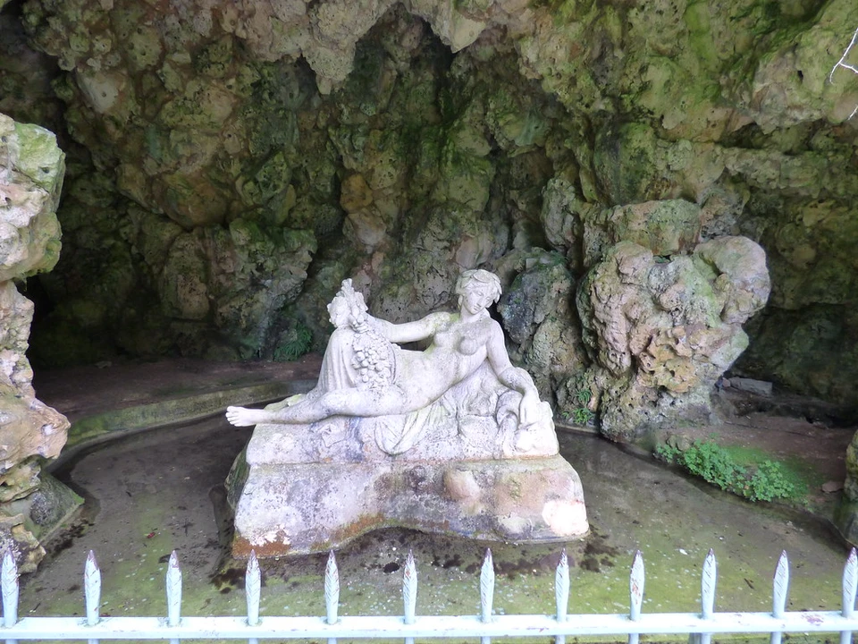 Künstliche steinerne Grotte an der Seinequelle mit liegender Nymphenfigur, Wasser und einem Gitter im Vordergrund; Aufnahme von 2014.