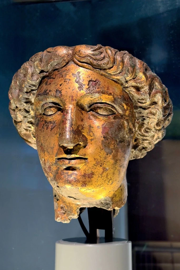 Vergoldeter Bronzekopf im Museum Bath, mit erhaltenen Locken, ungleichmäßig goldfarbener Oberfläche und abgebrochener Halskante auf einer modernen Halterung.