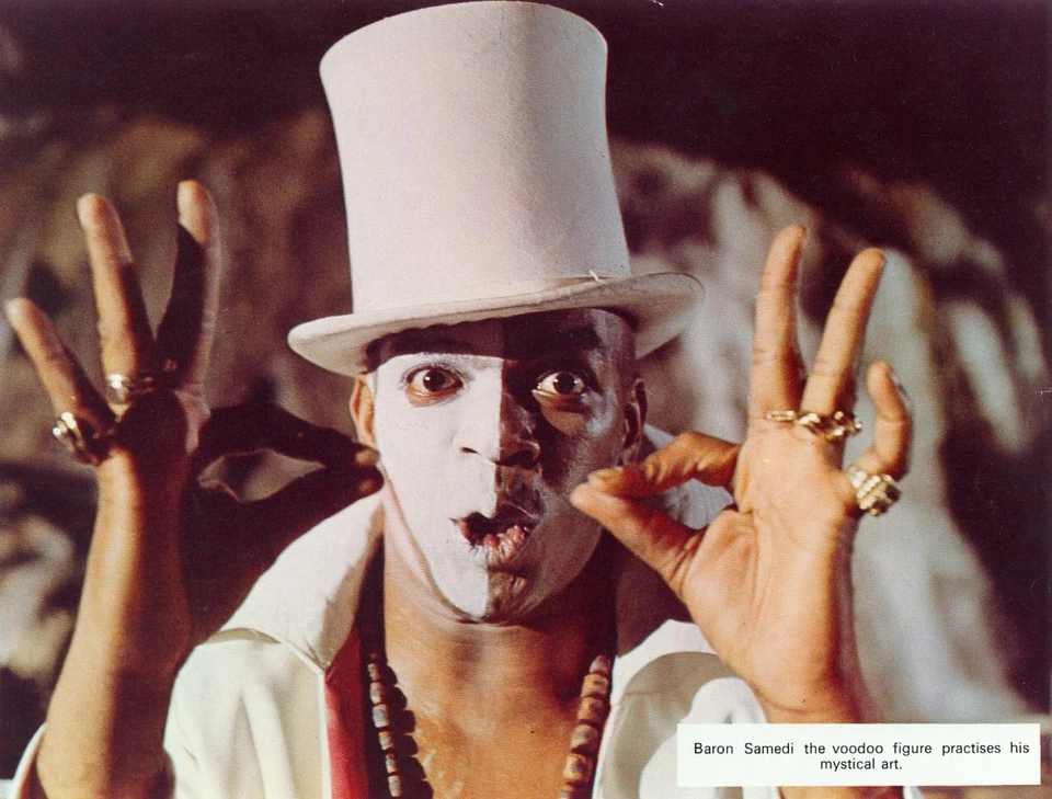Geoffrey Holder mit hohem weißem Zylinder, heller Gesichtsbemalung, dunklen Augenpartien und erhobenen Händen; die englische Bildzeile bleibt im Bild.